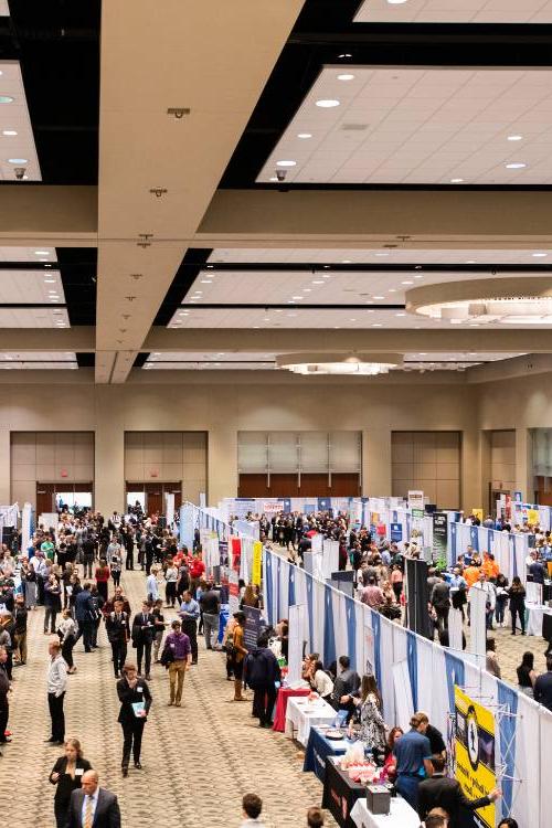 Large room with booths in multiple rows with many people walking down the aisles